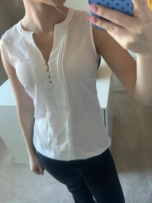 Vit ärmlös blus med knappar - Snygg vit ärmlös blus med dekorativa knappar framtill och diskreta sömdetaljer längs bröstet. Blusen har en lätt v-ringad halsringning och är tillverkad i ett mjukt, tunt material som känns skönt mot huden. Perfekt till jeans eller kjol för en fräsch look.