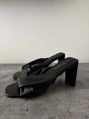 Svarta pumps med bred klack från Duffy - Säljer ett par mycket bekväma svarta klackar från Duffy med bred klack och trendig fyrkantig tå. Skorna har en vadderad, korsad rem i konstläder som ger en cool och modern look. Perfekta till sommaren! 