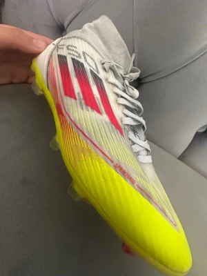 Adidas X Speedflow fotbollsskor gul/vit - Säljer ett par Adidas X Speedflow fotbollsskor med en snygg kombination av gul, vit och röda detaljer. Skorna har Sprintframe-yttersula för grymt grepp på planen och ovandel i lätt syntetmaterial. Perfekta för dig som vill ha snabbhet och stil på fotbollsplanen.