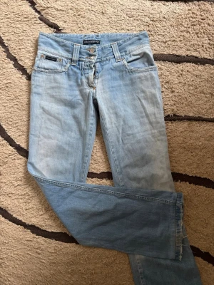Ljusblå jeans från Dolce & Gabbana - Snygga ljusblå jeans från Dolce & Gabbana med klassisk femficksdesign och raka ben. Jeansen har en cool tvättad look och detaljer som logotyp-patch bak samt metallknapp framtill, kedjan är sönder men kan får fixas av köparen och därför säljer jag de till ett väldigt bra pris