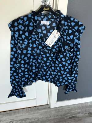 Blå hjärt-mönstrad blus Holly & Whyte - Snygg blus från Holly & Whyte i mörkblått med ljusblå hjärtan. Blusen har långa ärmar med volang vid ärmslut, v-ringning med knytband och lätt volang vid axlarna. Tillverkad i ett tunt, luftigt material som känns skönt mot huden.