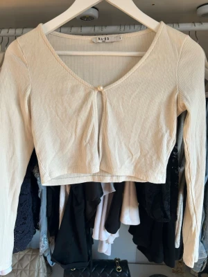 Ribbad croppad topp från NA-KD - Trendig ljusbeige ribbad croppad topp från NA-KD med lång ärm och v-ringning. Toppen har en liten pärldetalj framtill och är tillverkad i mjukt material som sitter snyggt på kroppen. Perfekt för att styla med höga jeans eller kjol.