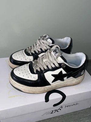 Svarta och vita Bape Sta sneakers - Säljer ett par klassiska Bape Sta sneakers i svart och vitt läder med den ikoniska stjärnan på sidan. Skorna har perforerad tå, vita snören och en chunky sula. Perfekta för dig som gillar streetwear och vill sticka ut med en unik look.
