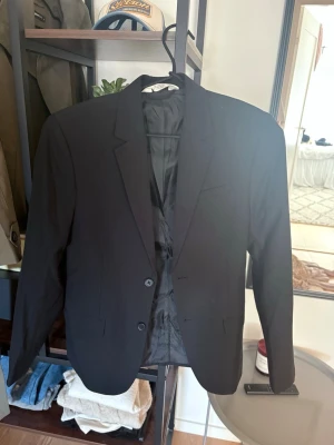 Svart klassisk kavaj med två knappar - Stilren svart kavaj med två knappar framtill och klassiska slag. Kavajen har en rak passform och är helfodrad med slät insida. Perfekt för en dressad look och enkel att matcha med olika outfits.