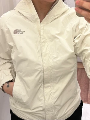 Vit regnjacka från The North Face - Snygg vit regnjacka från The North Face med huva och dragkedja framtill. Jackan har en clean look, diskret logga på bröstet och är tillverkad i lätt syntetmaterial som står emot regn. Perfekt för blöta dagar och enkel att matcha med allt.