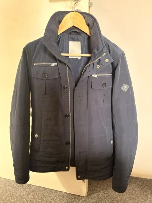 Mörkblå overshirt från J.Lindeberg - Snygg mörkblå pufferjacka från J.Lindeberg med hög krage, två bröstfickor med knappar och dragkedjor samt emblem på ärmen. Jackan har en stilren design och stängs med både dragkedja och knappar. Perfekt för kalla dagar.