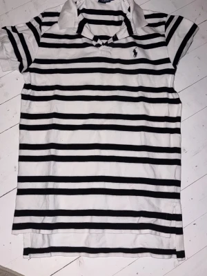 Randig pikétröja från Ralph Lauren - Klassisk vit pikétröja från Ralph Lauren med svarta horisontella ränder och liten broderad logga på bröstet. Tröjan har krage, korta ärmar och är gjord i mjuk bomull. Perfekt för en avslappnad men ändå stilren look.