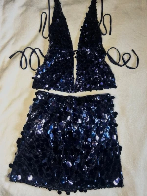Marinblå paljettopp med knytband och kjol set  - Snygg marinblå topp med djup v-ringning och knytband i sidorna och med matchande kjol. Setet är täckt av glittrande paljetter som ger en riktigt festlig look. Materialet är syntet och toppen är croppad med öppen rygg. Perfekt för dig som vill sticka ut.
