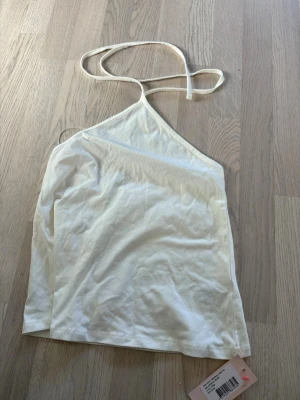 Vit halterneck topp från Nelly - Säljer en vit halterneck topp från Nelly med smala band som knyts i nacken. Toppen har en enkel och stilren design i mjuk bomull, perfekt för varma dagar. Modellen är loose fit och ärmlös för en avslappnad look. Strl S oanvänd 