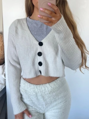 Kofta - Säljer en vit croppad kofta med ribbad struktur och fyra stora svarta knappar framtill. Koftan har v-ringning och långa ärmar, perfekt för en chill och trendig look. Råkat tvätta den med mörka kläder därav går den mot det grå hållet. Storlek S 🩷