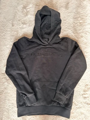 Svart hoodie från Peak Performance - Svart hoodie från Peak Performance med broderad logga på bröstet. Klassisk känguruficka framtill och ribbade muddar vid ärmar och nederkant. Tillverkad i mjuk bomull, perfekt för chill dagar eller när du vill ha en clean look.