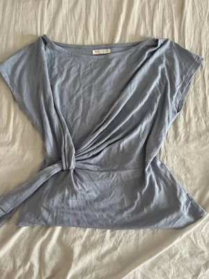 Ljusblå omlottopp från Karol&T - Snygg ljusblå omlottopp från RABALDER med korta ärmar och knytning i midjan. Tillverkad i mjukt, tunt tyg som ger en avslappnad och trendig look. Perfekt att matcha med jeans eller kjol för en cool stil.