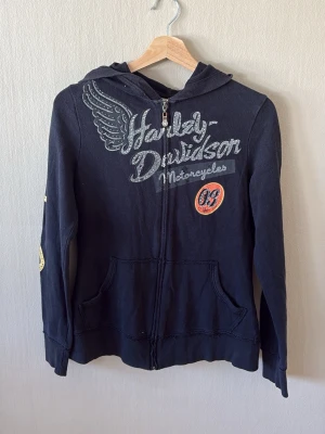 Y2K Svart Harley-Davidson hoodie med tryck - Säljer en svart hoodie från Harley-Davidson med dragkedja, huva och coolt tryck framtill med vinge och logga. Fickor på magen och patchdetalj med siffran 69. Tillverkad i 100% bomull med muddar vid ärmslut och nederkant. Perfekt för dig som gillar bikerstil.