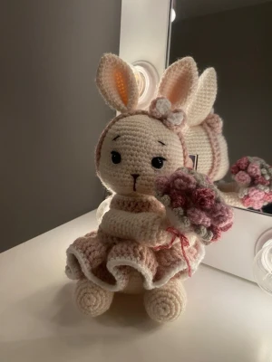 Virkat kaningosedjur med klänning - Handgjord virkad kanin i beige och rosa med stora öron, klänning med volang och en liten blombukett i handen. Kaninen har svarta broderade ögon och en rosett på huvudet. 