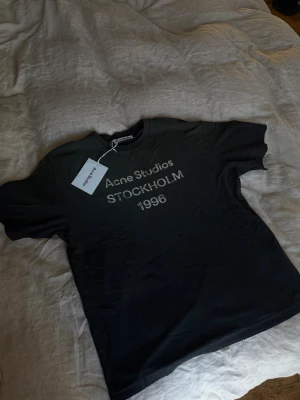 Svart Acne Studios t-shirt - Snygg svart t-shirt från Acne Studios med tryck 'Acne Studios STOCKHOLM 1996' på bröstet. Klassisk rund hals och korta ärmar. Tillverkad i mjuk bomull för en skön känsla. Perfekt för dig som gillar stilrena och trendiga plagg.