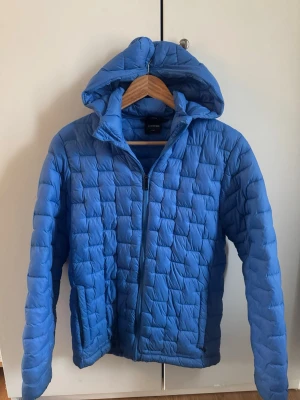 Blå dunjacka med huva från Everest - Snygg blå dunjacka från Everest med quiltat mönster och praktisk huva. Jackan har dragkedja framtill och två sidofickor med dragkedja. Perfekt för kalla vinterdagar och riktigt skön att bära. Klassisk modell som funkar till det mesta.