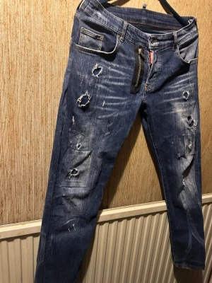 Dsq2 jeans  - Säljer ett par blå jeans med flera slitningar och hål på benen för en riktigt edgy look. Jeansen har klassisk femficksmodell, dragkedja och knapp i midjan. Perfekta för dig som gillar streetstyle och vill sticka ut. Midjamått 45cm,längden 105