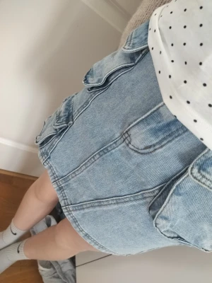 Jättefin jeanskjol - Snygg ljusblå jeanskjol från Yoyo S. med flera stora fickor framtill och baktill. Kjolen har en klassisk jeanslook med synliga sömmar, bälteshällor och knappstängning. Perfekt för dig som gillar praktiska detaljer och streetstil. STORLEK 10 ÅRINGAR