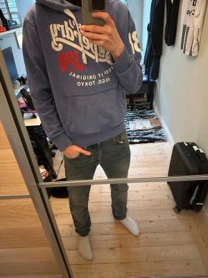 Blå Superdry hoodie med tryck - Säljer en blå hoodie från Superdry med stor vit och röd logga på bröstet och tryck på ena ärmen. Hoodien har huva med dragsko och en klassisk magficka. Riktigt bra skick utan defekter. Hör av er vid minsta fundering!