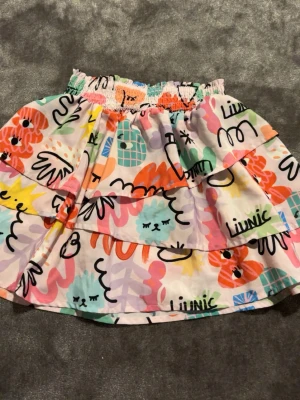 Färgglad kortkjol från H&M - Superfin kortkjol från H&M med färgglatt blommigt och grafiskt mönster i rosa, orange, blått, grönt och gult. Kjolen har volangdetaljer och resår i midjan för skön passform. Perfekt för dig som gillar lekfull stil!