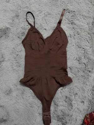 Brun ribbad body med smala band - Snygg brun body med ribbad struktur och smala axelband. Djup urringning framtill och knäppning i grenen. Perfekt att styla under skjortor eller tröjor för en trendig look. Materialet känns mjukt och stretchigt mot huden.