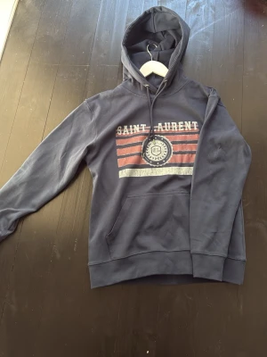 Mörkblå Saint Laurent hoodie - Snygg mörkblå hoodie från Saint Laurent med tryck framtill i vitt och rött. perfekt för en avslappnad och trendig look.