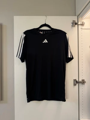 Svart träningströja med vita detaljer - Snygg svart träningströja från Adidas med klassiska vita ränder på axlarna och Adidas-logga på bröstet. Tillverkad i ett lätt och funktionellt material som passar perfekt till träning eller sportaktiviteter. Kortärmad och med rund hals.💙 aldrig använd!