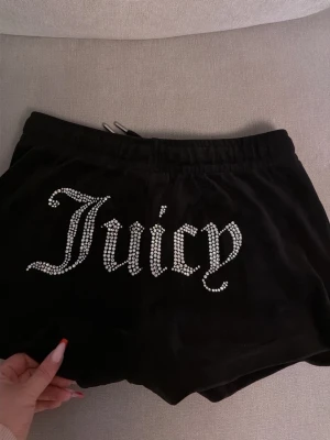 Svarta Juicy Couture shorts i velour - Säljer ett par svarta velourshorts från Juicy Couture med glittrande strasstenar som stavar 'Juicy' på baksidan och ett litet J på framsidan. Shortsen har dragsko i midjan och är perfekta för en avslappnad och trendig look.  Pris kan diskuteras 