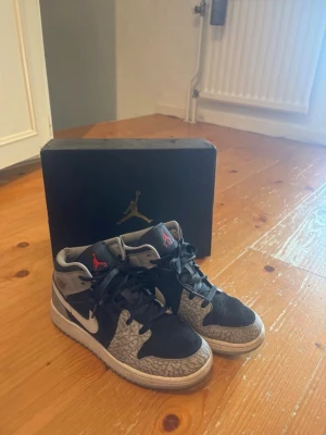 Air Jordan 1 Mid Elephant Print - Säljer ett par Air Jordan 1 Mid med ikoniskt elephant print i grått och svart. Skorna har svart och grå mocka, vita swoosh-loggor och röda detaljer på plösen. Klassisk Jordan-logga på sidan och bekväm sula. Perfekta för dig som gillar streetwear och sneakers med unik design. Lite slitna inuti men utanpå är de i riktigt bra skick