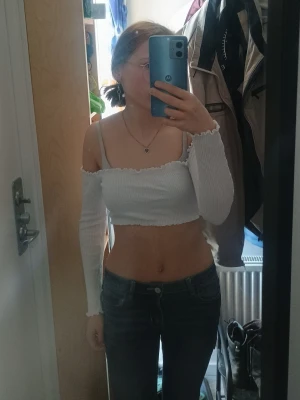 Vit croppad offshoulder topp - Säljer en vit croppad offshoulder topp med långa ärmar och ribbad struktur. Toppen har en söt volangkant upptill och nedtill och sitter tight över kroppen. Perfekt för dig som gillar trendiga och feminina plagg.