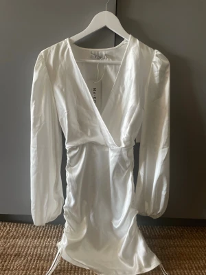 Vit omlottklänning från NA-KD - Säljer en vit kort omlottklänning från NA-KD i glansigt material med långa puffärmar och snörning i sidan. Klänningen har v-ringning och en elegant, figurnära passform. Perfekt för dig som vill ha en stilren och trendig look. Passar super till studenten eller skolavslutning 