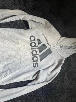 Grå hoodie från Adidas med logga - Snygg grå hoodie från Adidas med stor svart logga på bröstet. Klassisk känguruficka framtill och justerbar huva med dragsko. Tillverkad i mjukt sweatshirtmaterial som är skönt mot huden. Perfekt för en avslappnad och sportig stil.