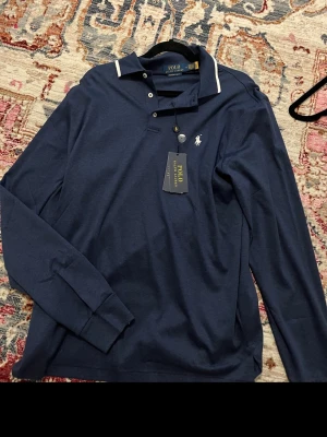 Marinblå långärmad skjorta Polo Ralph Lauren - Snygg marinblå långärmad skjorta från Polo Ralph Lauren med klassisk krage, knappar framtill och vit broderad logga på bröstet. Skjortan har en stilren look med diskreta vita detaljer vid kragen och är tillverkad i mjuk bomull.