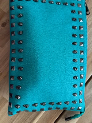 Turkos kuvertväska med nitar från Tiamo - Snygg turkos kuvertväska från Tiamo med silverfärgade nitar och dödskalle-detaljer runt kanterna. Väskan har dragkedja och insidan är fodrad med leopardmönster. Perfekt för dig som gillar att sticka ut och vill ha en edgy accessoar.