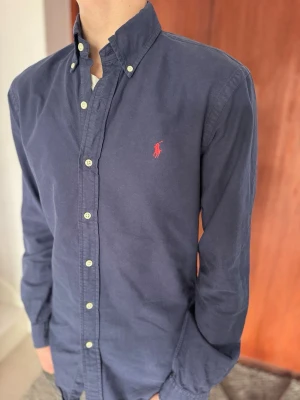 Mörkblå skjorta från Ralph Lauren - Säljer en stilren marinblå långärmad oxfordskjorta från Polo Ralph Lauren. Det är en Slim Fit-modell i herrstorlek Small, vilket ger en snygg och modern passform.  • Märke: Polo Ralph Lauren • Modell: Oxford Shirt • Passform: Slim Fit • Storlek: Small (Herr) • Färg: Marinblå • Nypris: ca 1 400 kr • Skick: Bra begagnat skick 