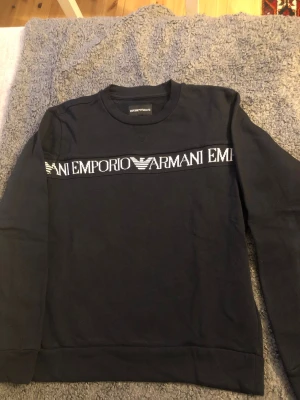 Svart sweatshirt från Emporio Armani - Snygg svart sweatshirt från Emporio Armani med vit logga och text tvärs över bröstet. Klassisk rund halsringning och långa ärmar. Perfekt för dig som gillar stilrena och exklusiva plagg med tydlig branding.passar till 11 år, som ny