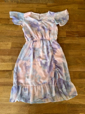 Pastellfärgad omlottklänning - Söt långklänning i pastellfärger med ljuslila, blå och rosa toner. Klänningen har korta, fladdriga ärmar, omlottdetalj med snörning i sidan och volang längst ner. Knäpps med knapp i nacken och har ett luftigt, lätt material som passar perfekt till varma dagar.