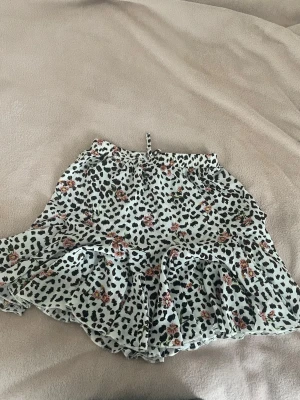 Leopardmönstrad kortkjol med volang - Supersöt kortkjol med leopardmönster och små blommor i rosa och rött. Kjolen har volangkant och en söt knytning i midjan. Materialet känns lätt och flowy.