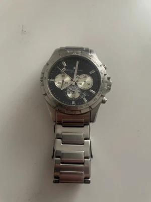 Jacques Lemans silverfärgad klocka - Stilren klocka från Jacques Lemans i silverfärgat metallarmband och boett. Urtavlan är svart med tre mindre kronografer och vita detaljer. Klockan har en sportig look med markerad bezel och robust länkarmband.