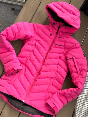 Rosa dunjacka från Peak Performance - Snygg och varm rosa dunjacka från Peak Performance med huva och dragkedja. Jackan har flera fickor, justerbar huva och klassisk quiltad design. Perfekt för kalla vinterdagar och riktigt skön att bära. Fint skick. Använd endast 2 gånger. Nypris ca 3500 kr . Storlek XS