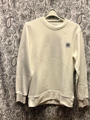 Vit sweatshirt från Stone Island  - Stilren vit sweatshirt från Stone Island med rund halsringning och diskret logopatch på bröstet. Tröjan har långa ärmar, ribbade muddar och är gjord i mjukt bomullsmaterial. Perfekt för en clean och avslappnad look. Finns i flera storlekar. 