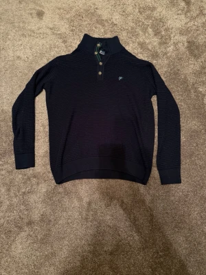 Mörkblå stickad tröja Denim Culture - Mörkblå stickad tröja från Denim Culture med krage och knappar framtill. Tröjan har diskret strukturmönster, ribbade muddar och gröna ränder på insidan av kragen. Liten broderad logga på bröstet. Perfekt för dig som gillar klassisk och stilren look. Storlek M