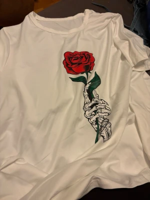 Vit t-shirt med röd ros och skelett - Snygg vit t-shirt med tryck av en röd ros som hålls av en skelett-hand. T-shirten har rund hals och korta ärmar. Perfekt för dig som gillar edgy och unika motiv. Materialet känns mjukt och skönt mot huden.