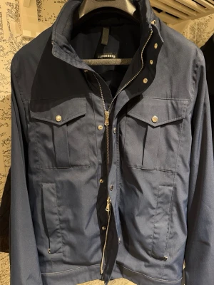 Mörkblå jacka från J.Lindeberg - Stilren mörkblå jacka från J.Lindeberg med två bröstfickor med lock och tryckknappar, dragkedja och knappstängning framtill. Jackan har en hög krage och är tillverkad i ett tåligt tyg med diskret logomärke på ärmen.