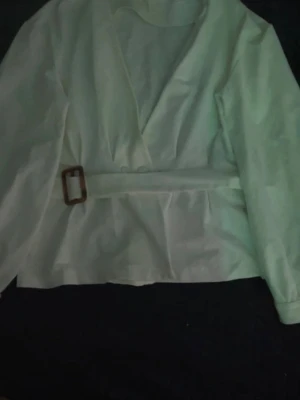 Vit omlottblus med bälte - Stilren vit omlottblus med lång ärm och markerad midja tack vare ett tygbälte med fyrkantigt spänne i trälook. Blusen har v-ringning och är gjord i ett lätt, vävt material som ger en snygg siluett.