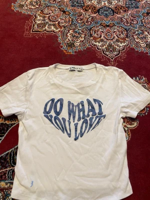 Vit t-shirt med blå texttryck - Vit t-shirt från Pull&Bear med blått texttryck i form av ett hjärta där det står 'DO WHAT YOU LOVE'. Klassisk rund halsringning och korta ärmar. Tillverkad i mjuk bomull för en skön känsla.