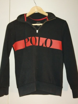 Svart tröja från Polo Ralph Lauren - Svart tröja från Polo Ralph Lauren med stor röd POLO-logga över bröstet. Tröjan har även en liten polo logga broderad på ena armen. Tröjan är väldigt sparsamt använd. Tröjan är en klassisk half-zip och är perfekt för dig som gillar att ha en avslappnad samt snygg stil.