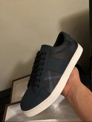 Mörkblå Burberry sneakers - Snygga mörkblå sneakers från Burberry med klassiskt rutmönster på sidan och vita sulor. Skorna har lågt skaft, svarta skosnören och detaljer i läder och textil. Perfekta för dig som vill ha en stilren och exklusiv look.