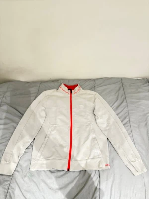 Vit ziptröja från BOSS med röd dragkedja - Säljer en stilren vit ziptröja från BOSS med röd dragkedja och detaljer. Tröjan har hög krage med BOSS-logga i rött runtom och diskret broderad logga på bröstet. Perfekt för dig som gillar sportig och clean stil.