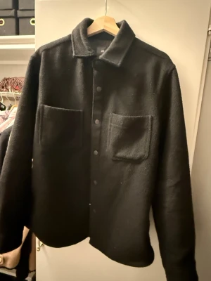 Svart overshirt jacka - Passar MStilren svart overshirt med klassisk krage och två stora bröstfickor. Jackan har tryckknappar framtill och är tillverkad i ett mjukt, lite tjockare tyg som ger en avslappnad look. Perfekt att slänga över en hoodie eller t-shirt för en cool lager-på-lager-stil.